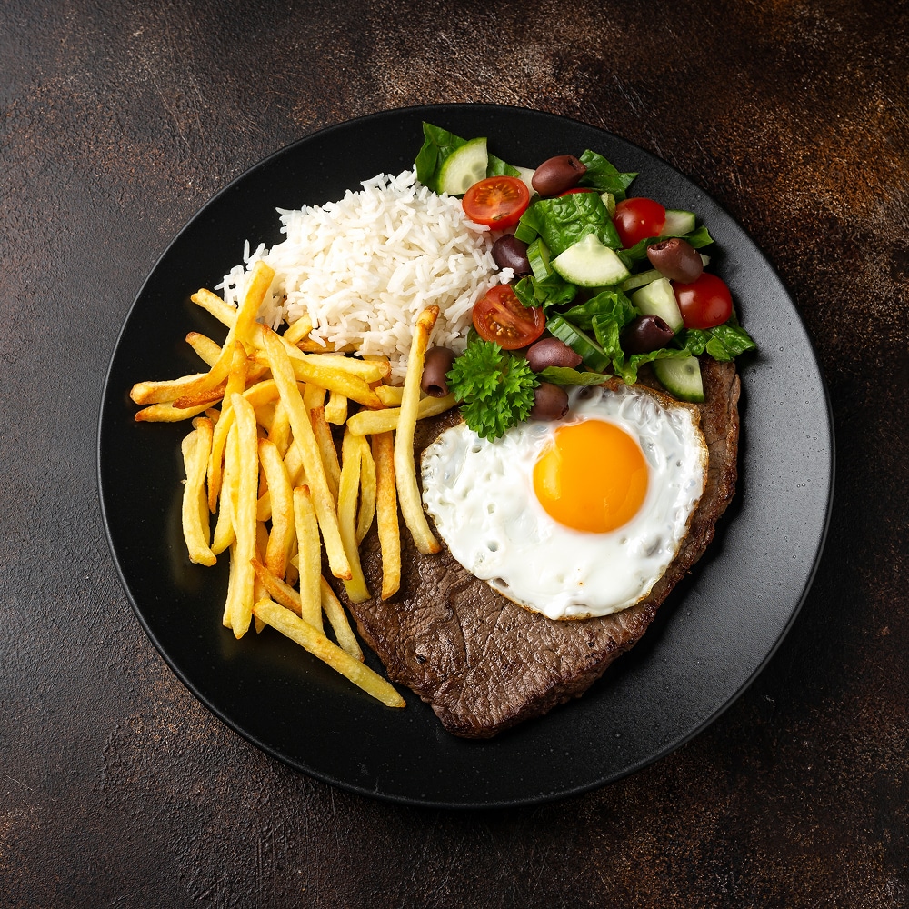 Bistec (steak) à l'œuf, riz et frites dorées sur assiette noire. Accompagné d'une salade de tomates, concombres et olives.