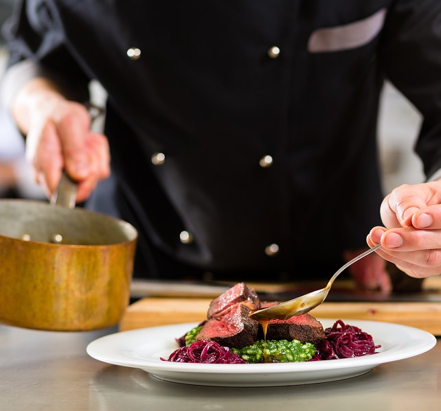 Dressage gastronomique : Chef nappe de sauce une viande rouge. Chef en veste noire nappe de sauce une pièce de viande rouge (saignante), accompagnée de chou rouge et purée verte, dans une cuisine pro.